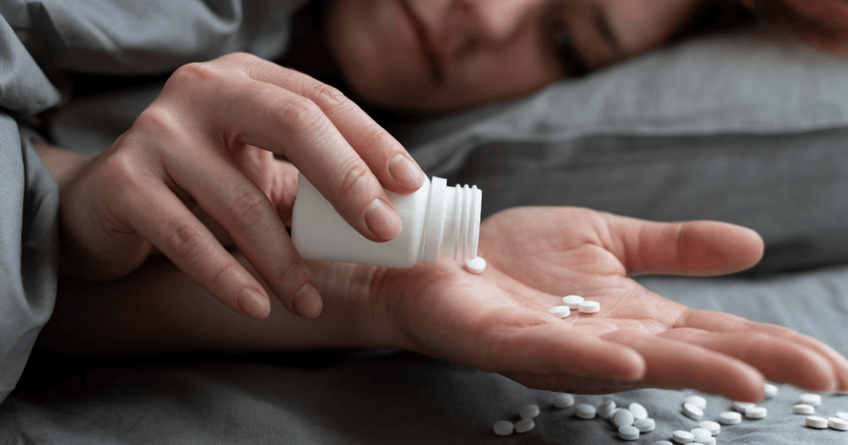 Imagem de uma mulher deitada na cama, com um pote de medicamentos sobre a sua mão.