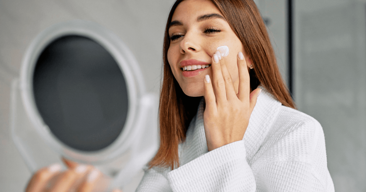 Imagem de uma mulher passando algum creme de proteção em seu rosto enquanto olha para o espelho.