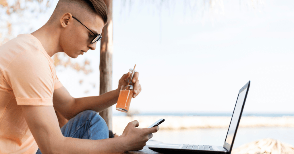 Imagem de um homem com seu celular e Notebook em frente a praia, mostrando a essência sobre o que é nômade digital.