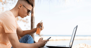 Imagem de um homem com seu celular e Notebook em frente a praia, mostrando a essência sobre o que é nômade digital.